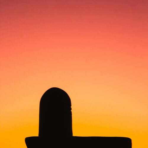 Silhouette of a Shiva Lingam against an orange and pink sunset sky in India.